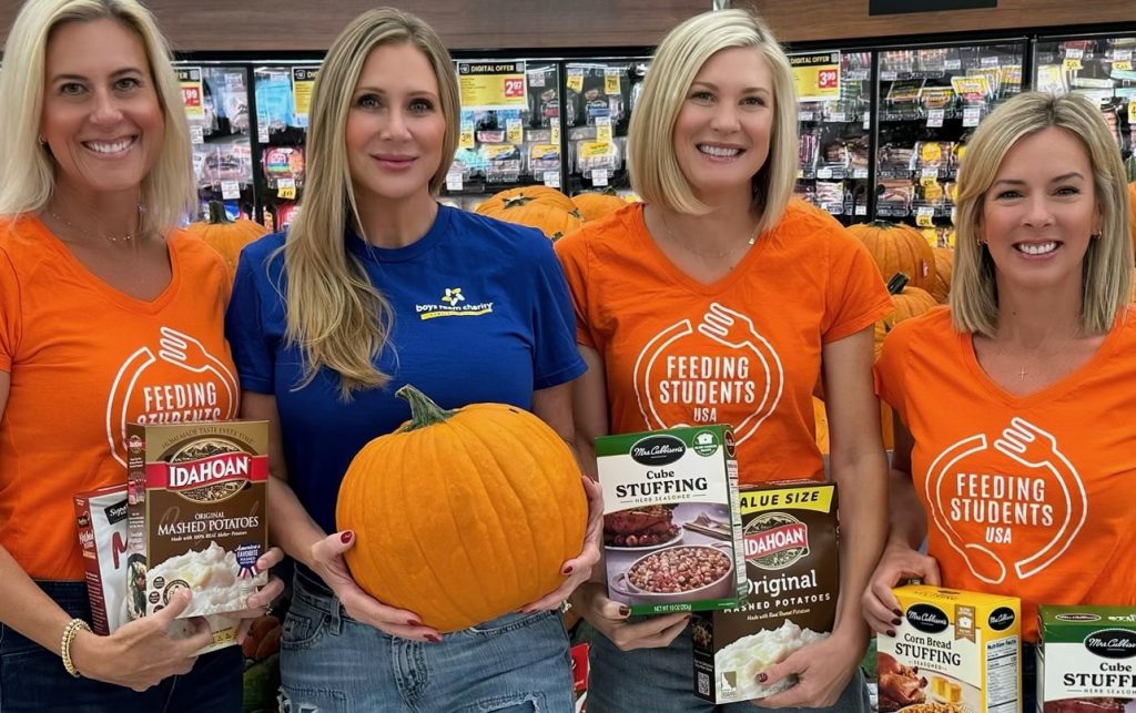 Four women, three in orange Feeding Students logo shirts, one in blue, are standing side-by-side in a grocery store holding up boxes of mashed potatoes and stuffing and a pumpkin.